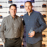 Two men holding a sailboat shaped award. The award is business of the year and then man on the left is Greg James and on the right is Jared DeWolf.