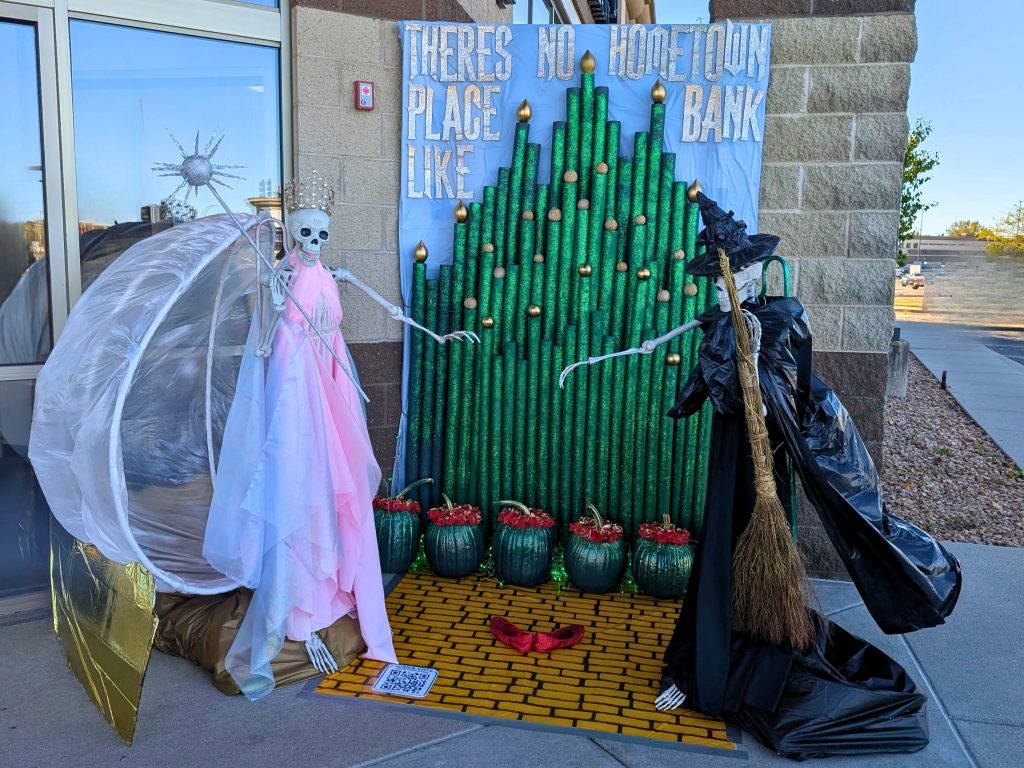 Two skeletons dressed in a pink dress and one in a black dress and black hat standing in front of a glittering green Emerald City.