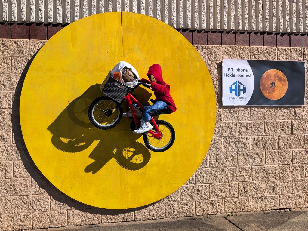 A scarecrow on a bike with an E.T. doll in the bike basket. The bike is riding across a full moon.