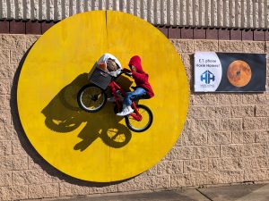 A scarecrow on a bike with an E.T. doll in the bike basket. The bike is riding across a full moon.