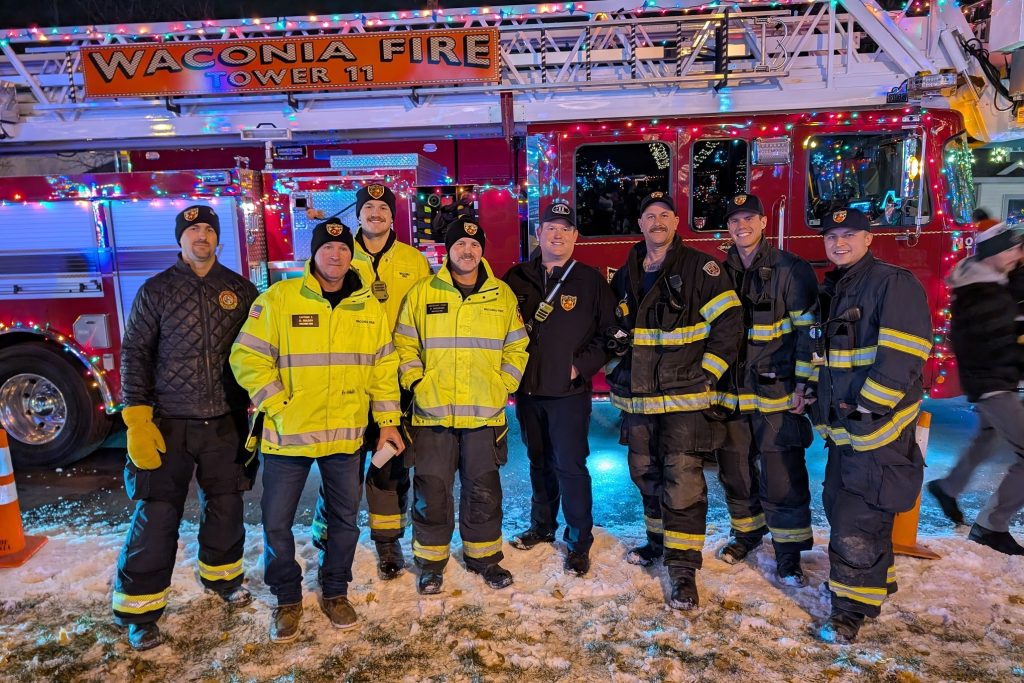 A group of firefighters standing in front of the lit up fire truck