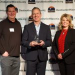 Three people posing with middle person holding award trophy