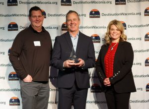 Three people posing with middle person holding award trophy