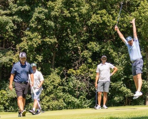 four guys on a golf putting green with one jumping in the air to celebrate